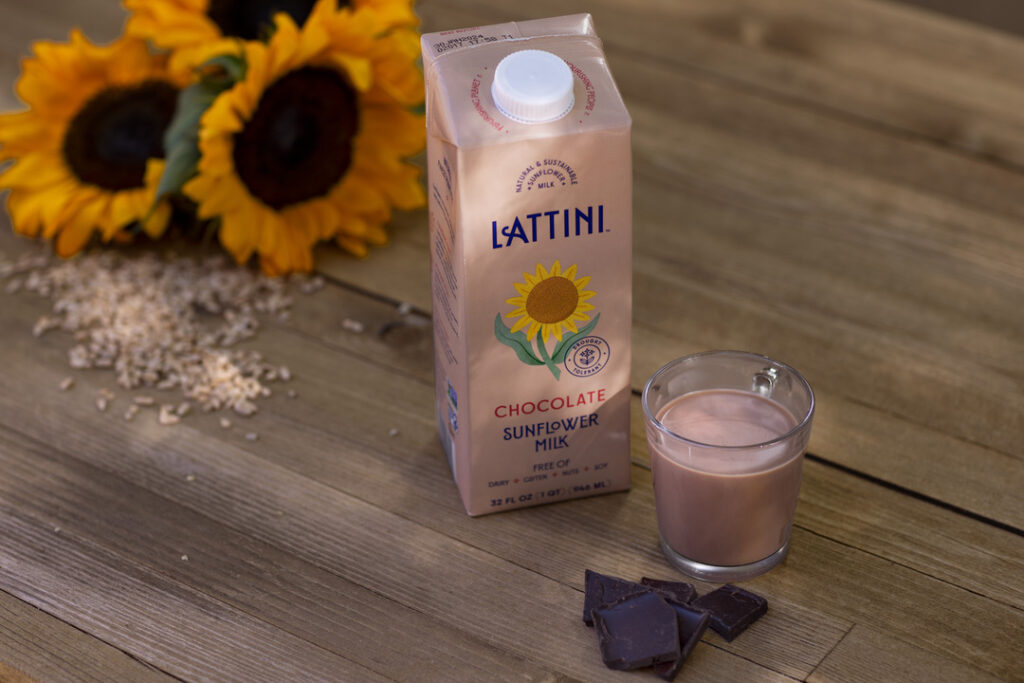 A carton and glass of the healthiest non-dairy milk sits on a wood counter next to oats, squares of chocolate, and sunflowers